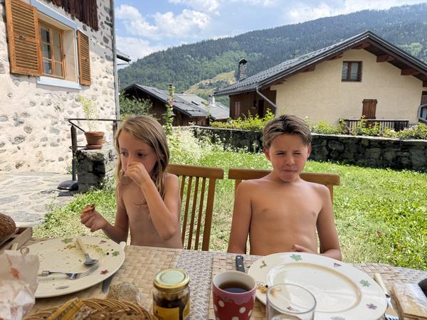 Two children are enjoying a sunny outdoor meal at Chemin de Fontaine Noire, Les Allues, with mountains and traditional houses in the background